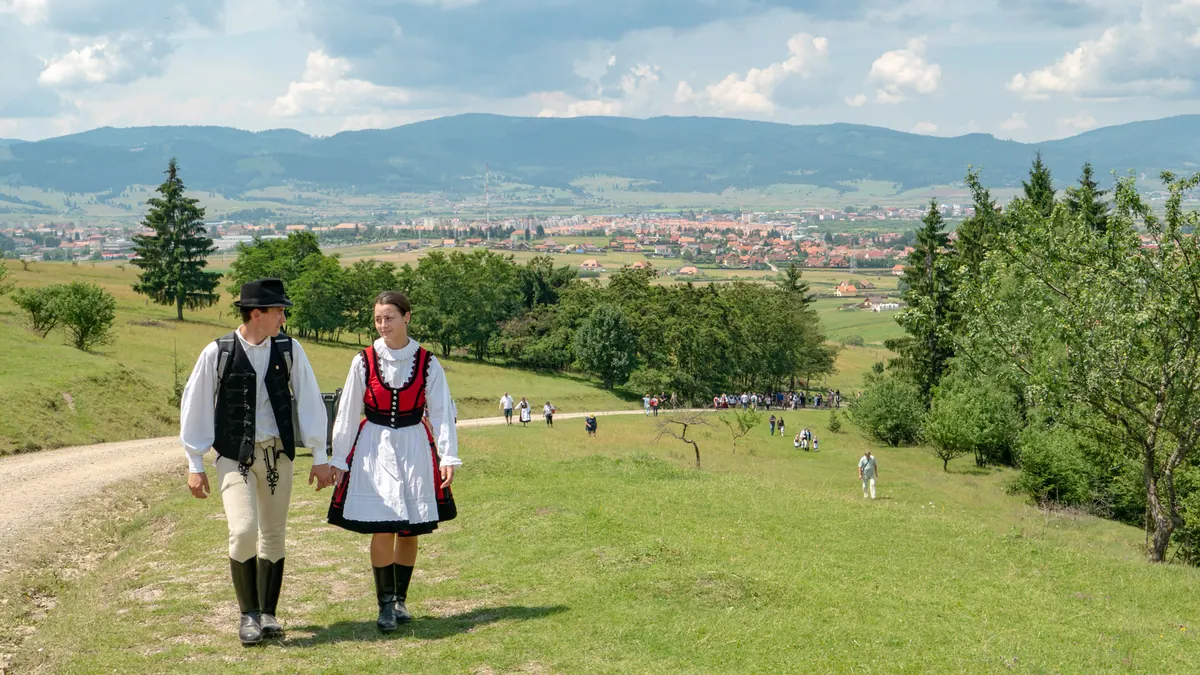 A couple in traditional Székely folk dress walking a hillside path above a valley town, Transylvania