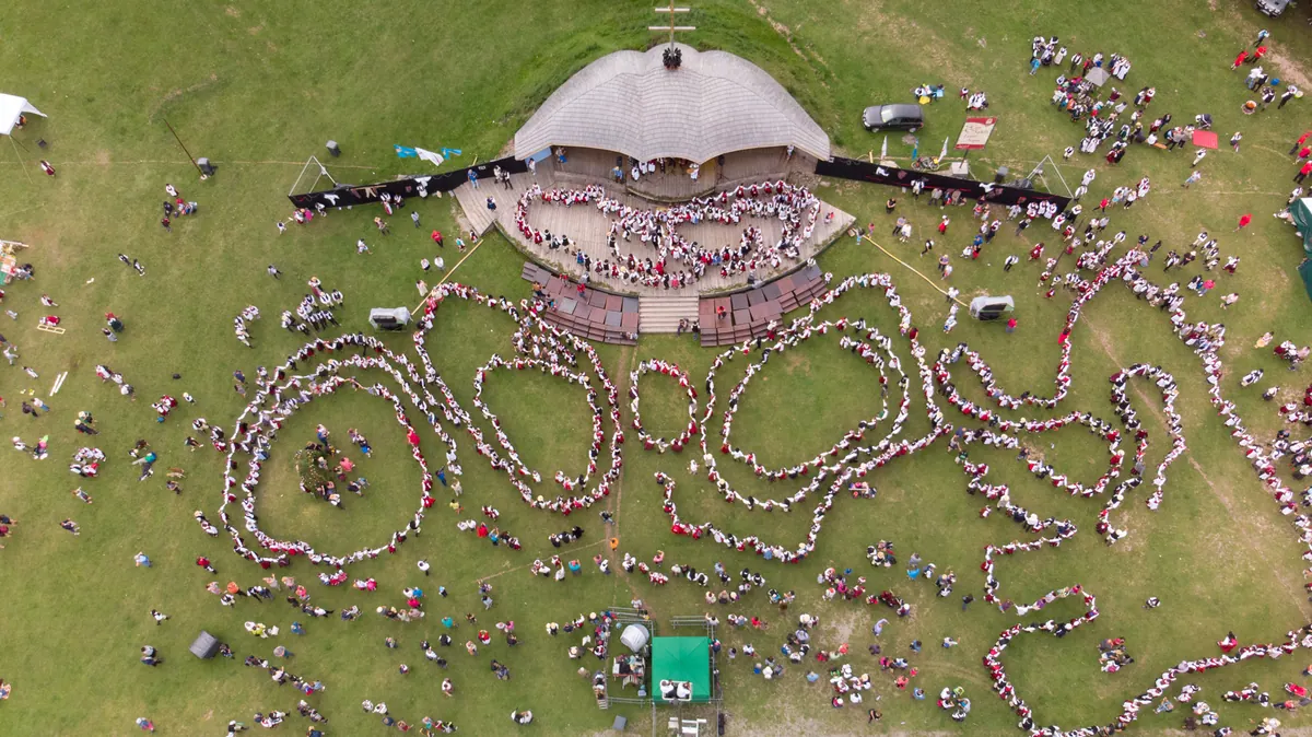 An aerial view of hundreds of dancers in red and white folk costume winding into spirals across a mountain meadow