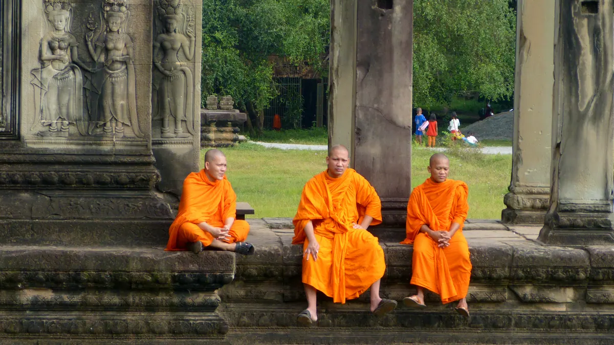 Three Buddhist monks in vivid orange robes resting among the stones of Angkor
