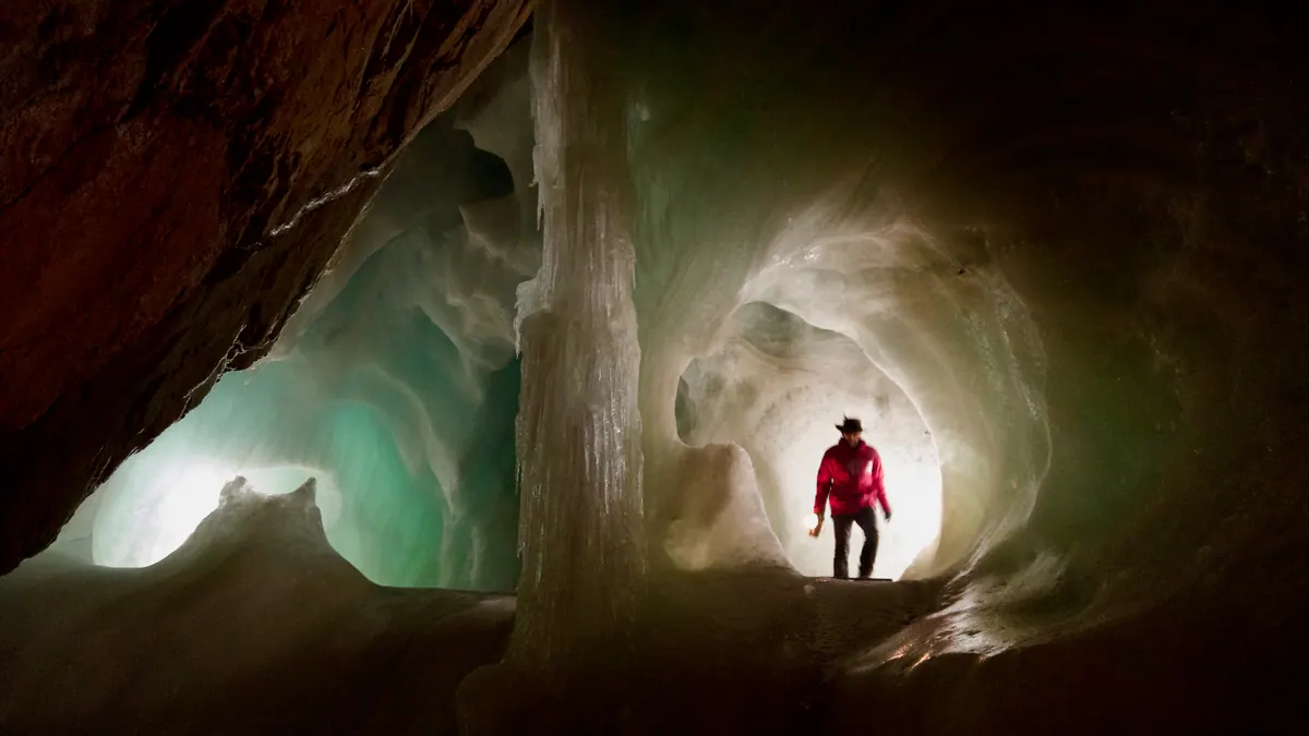 A lone figure in a red jacket standing in the glowing, sculpted passage of a vast ice cave