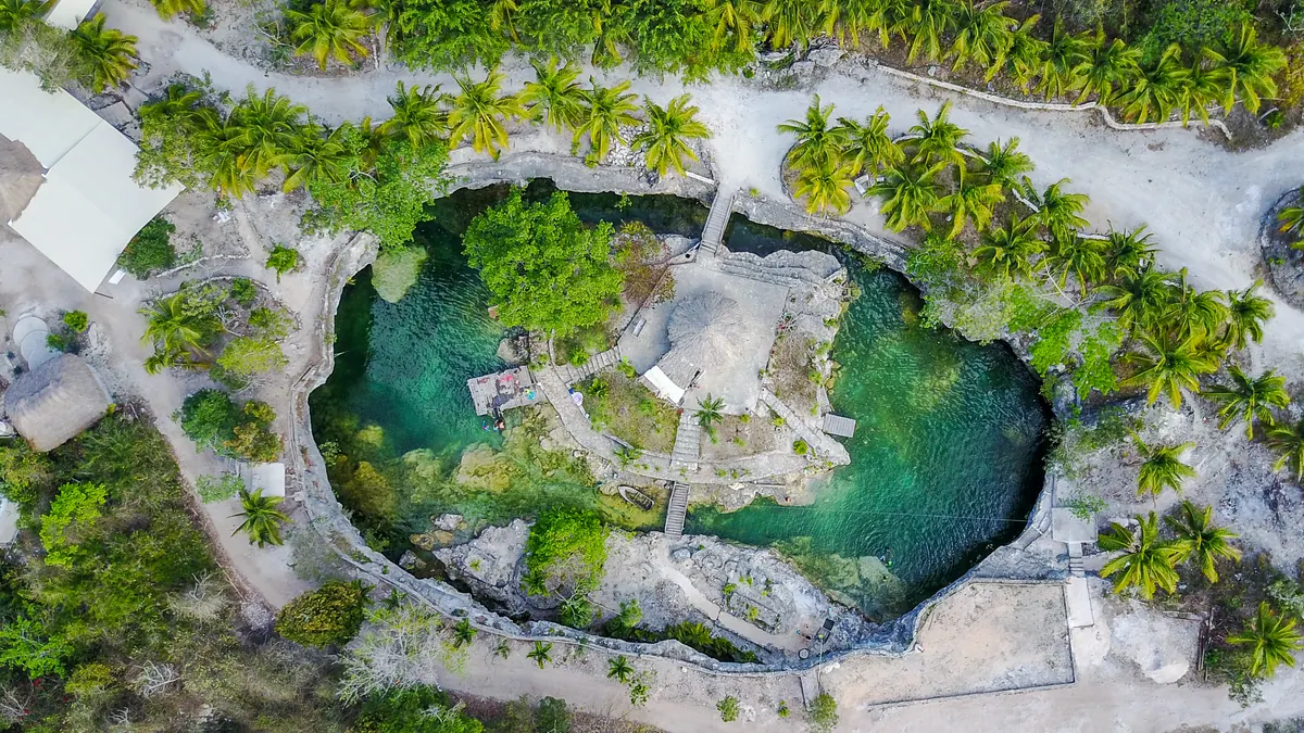 An aerial view looking straight down onto a turquoise cenote ringed by palms and limestone paths