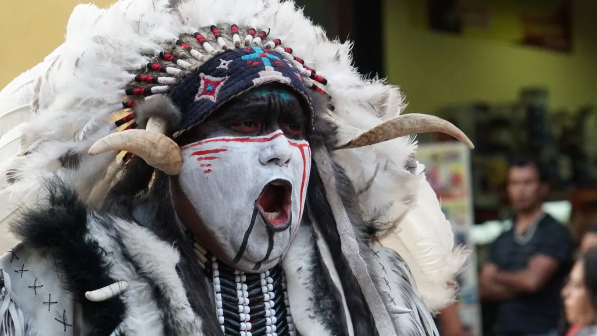 A dancer in a white feather headdress and face paint calling out during a street procession