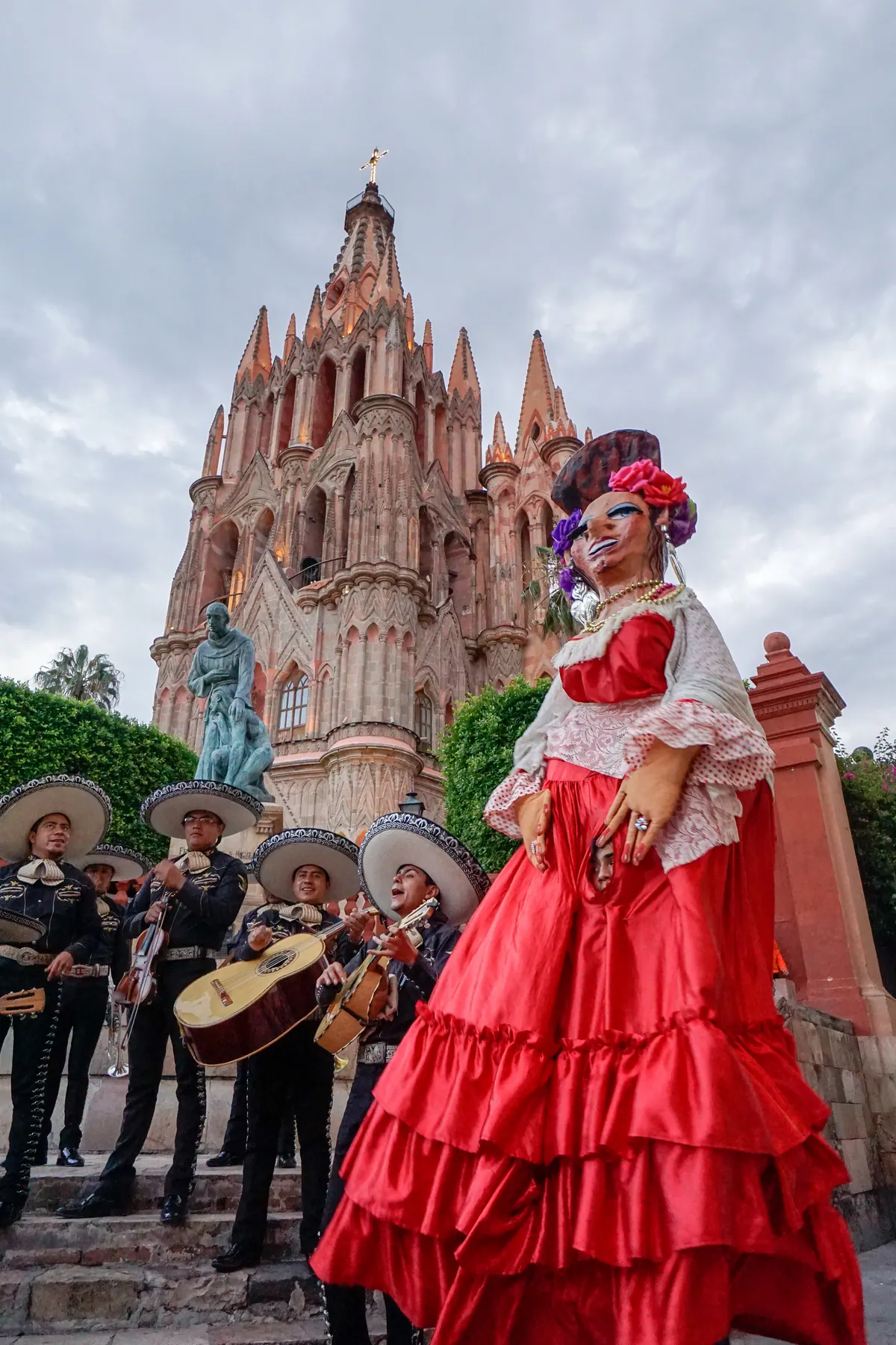 A towering mojiganga puppet in a red dress standing before the Parroquia while mariachis play on the steps below